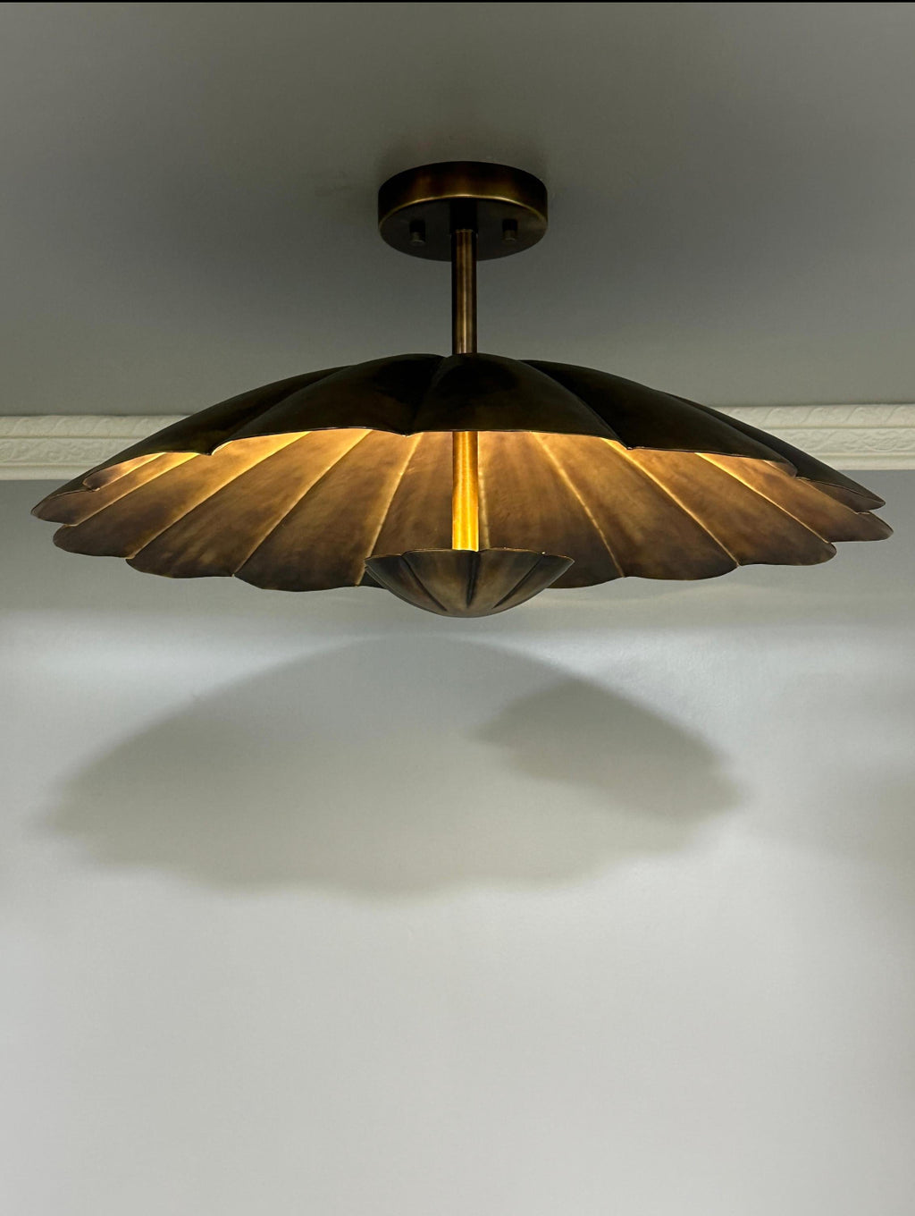 A modern semi-flush mount ceiling light fixture with a bronze metal shade in a scalloped, petal-like design, casting a warm golden glow downward against a plain white wall.