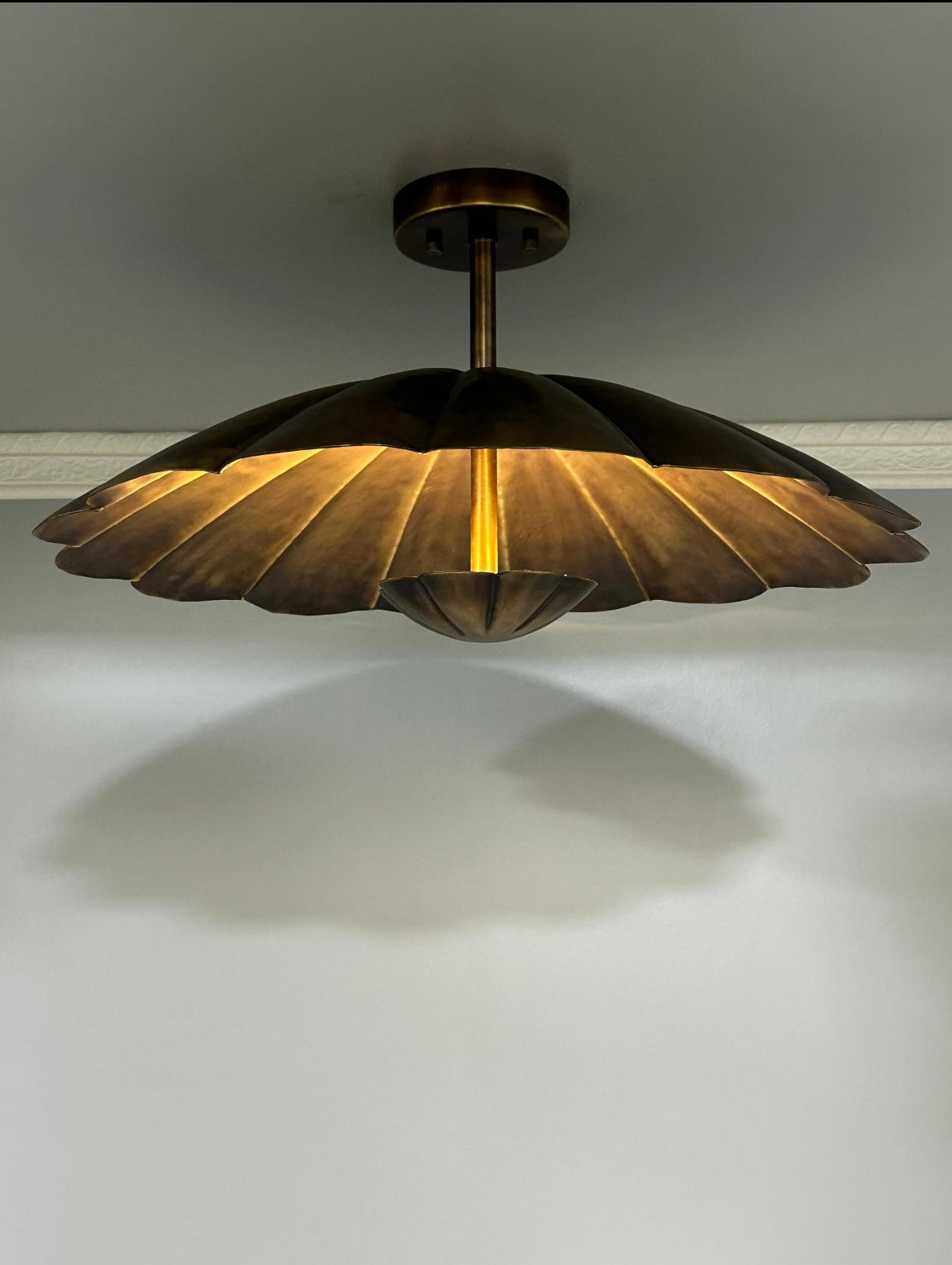 A modern semi-flush mount ceiling light fixture with a bronze metal shade in a scalloped, petal-like design, casting a warm golden glow downward against a plain white wall.
