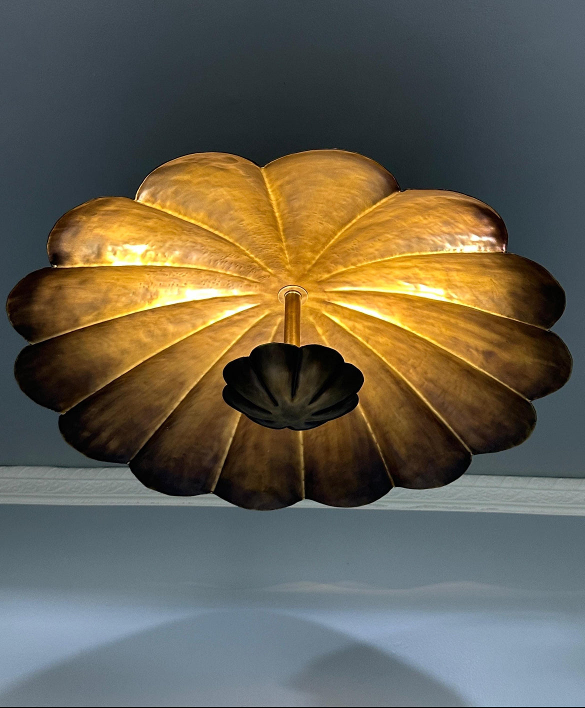 A sculptural, fan-shaped ceiling light fixture made of metallic gold-toned petals, shown illuminated against a dark gray wall.