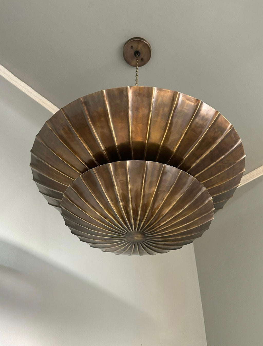 A large, tiered ceiling light fixture made of metallic bronze-toned metal with a pleated, fan-like design, suspended from a ceiling.