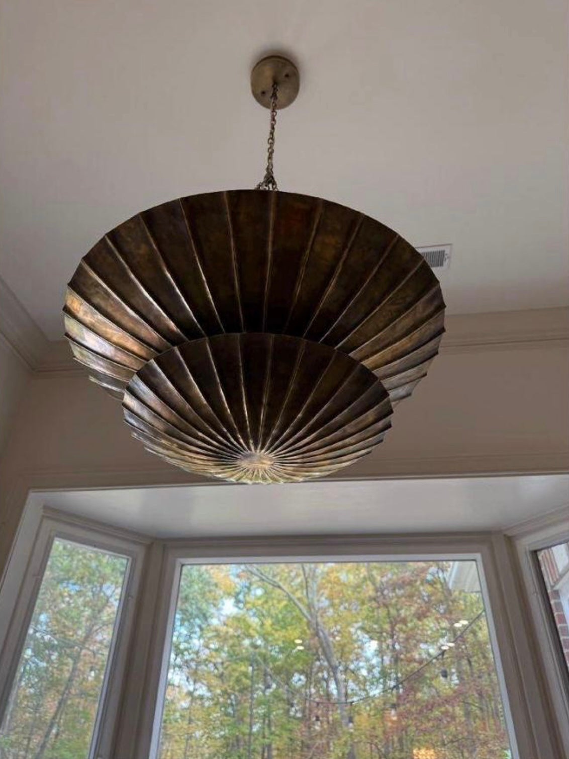 A large, tiered fan-shaped pendant chandelier with a distressed bronze finish, suspended from the ceiling in a home interior with bay windows.