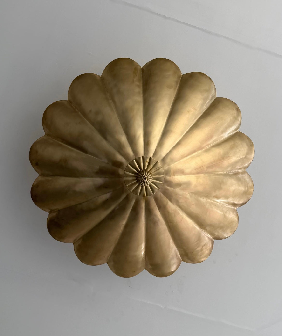 A round, scalloped brass wall sconce or decorative ceiling medallion with a textured central hub, shown from a top-down perspective against a plain light gray wall.