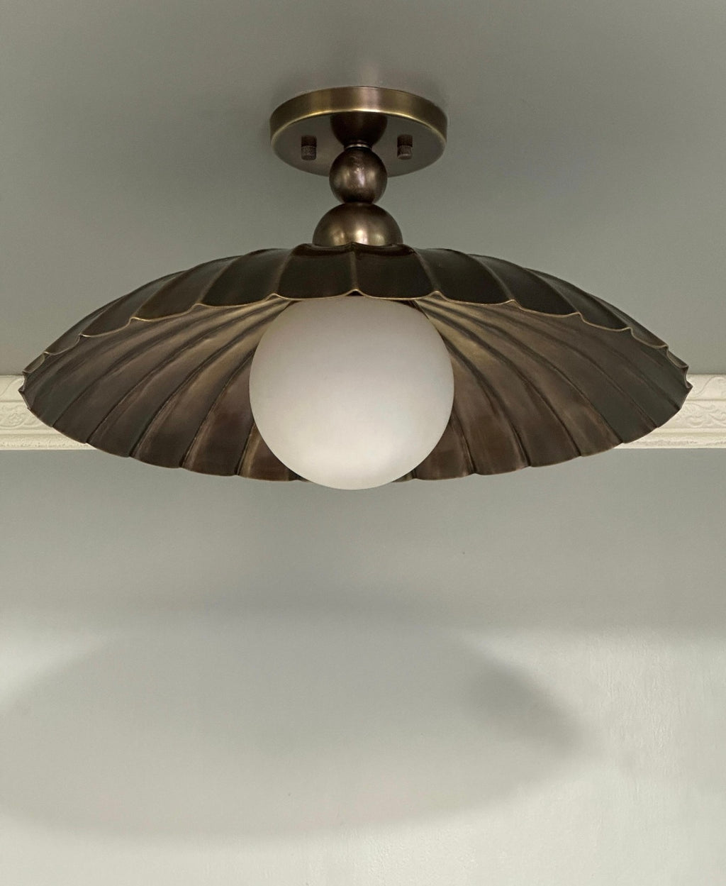 A vintage-style ceiling light fixture with a bronze-toned metal shade in a scalloped or fan-like shape surrounding a frosted white globe bulb, shown mounted on a ceiling.