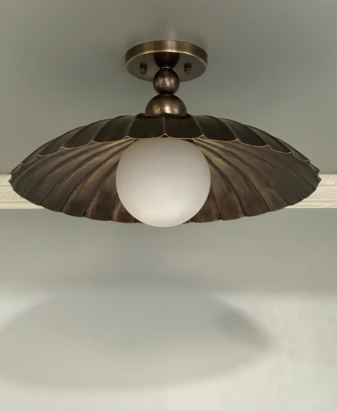 A vintage-style ceiling light fixture with a bronze-toned metal shade in a scalloped or fan-like shape surrounding a frosted white globe bulb, shown mounted on a ceiling.