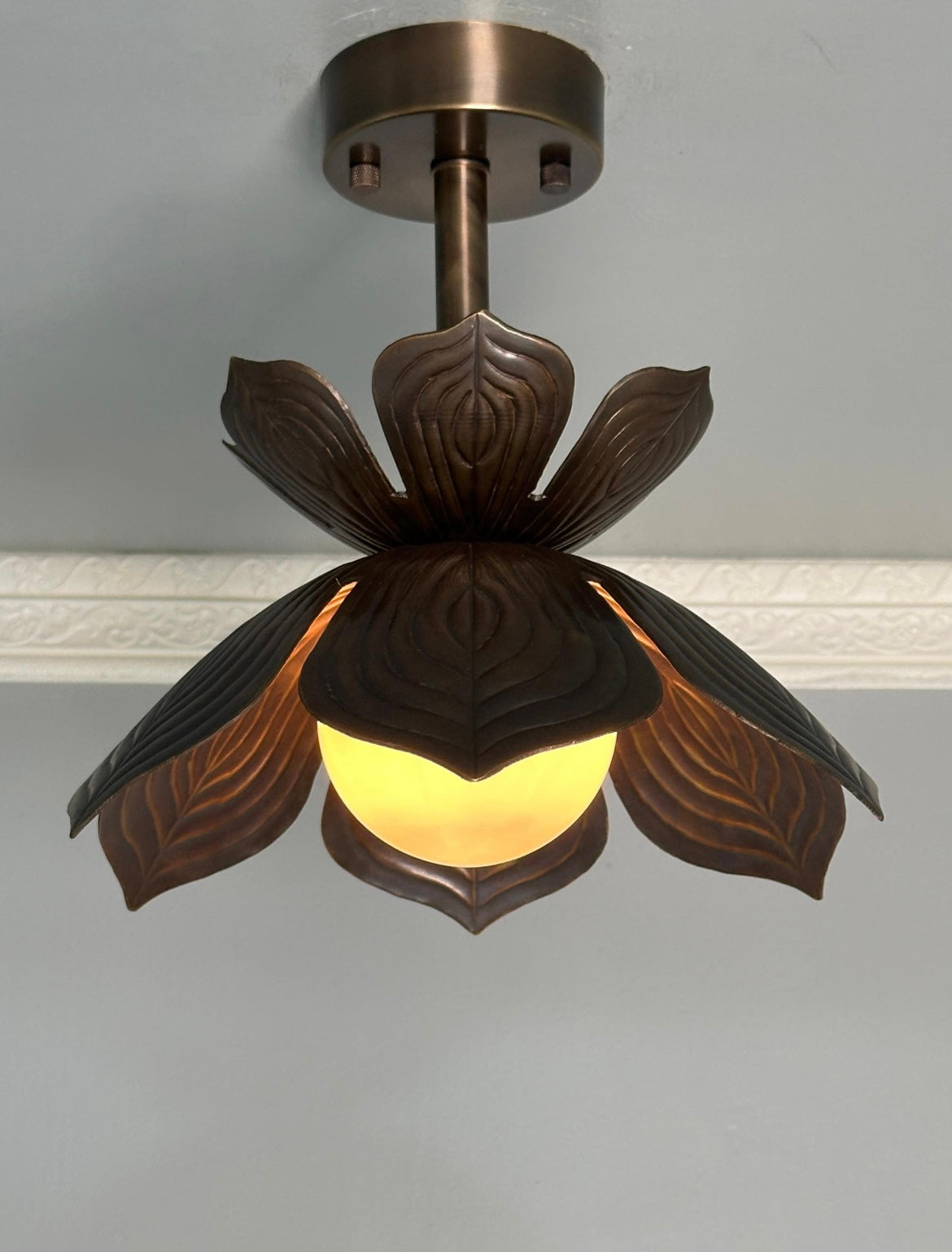 A ceiling-mounted pendant light fixture with a dark bronze metal frame shaped like a lotus flower, featuring carved leaf-like petals and a glowing warm yellow globe bulb.