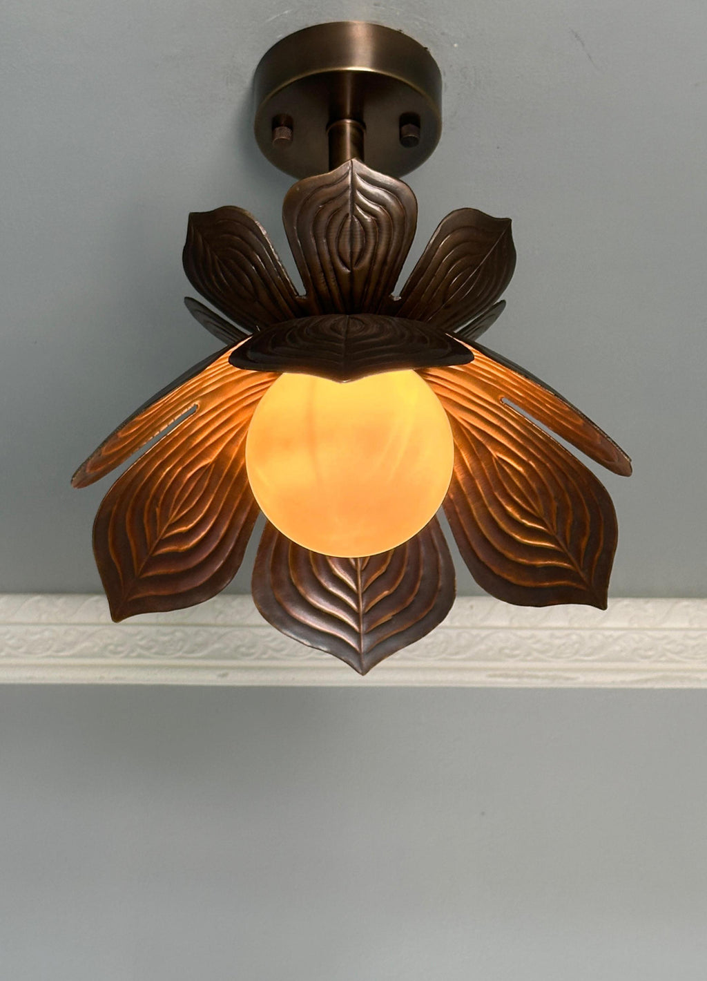 A decorative ceiling light fixture with a dark bronze metal leaf-like shade and a glowing warm yellow glass globe, mounted against a light gray wall.