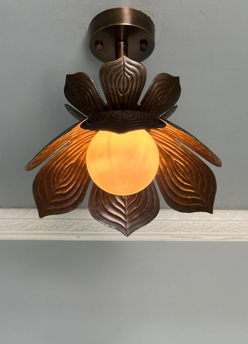 A decorative ceiling light fixture with a dark bronze metal leaf-like shade and a glowing warm yellow glass globe, mounted against a light gray wall.
